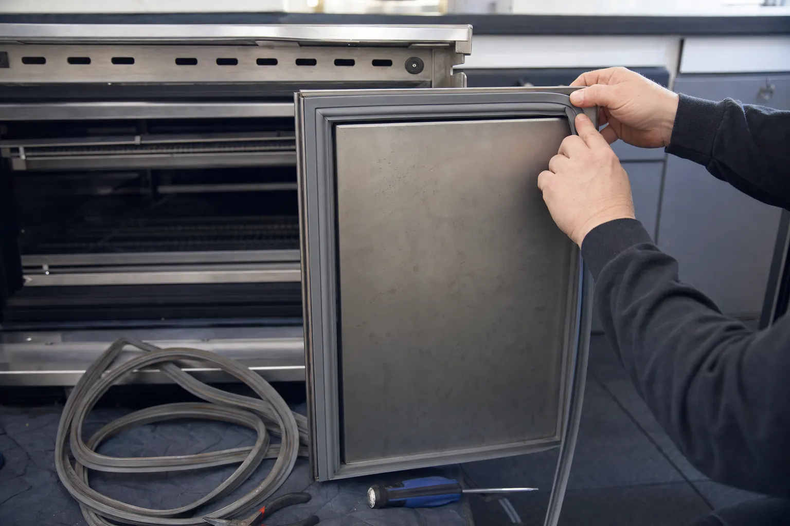 A replacement door gasket being pressed into the channel of a stainless undercounter door, the old one coiled nearby