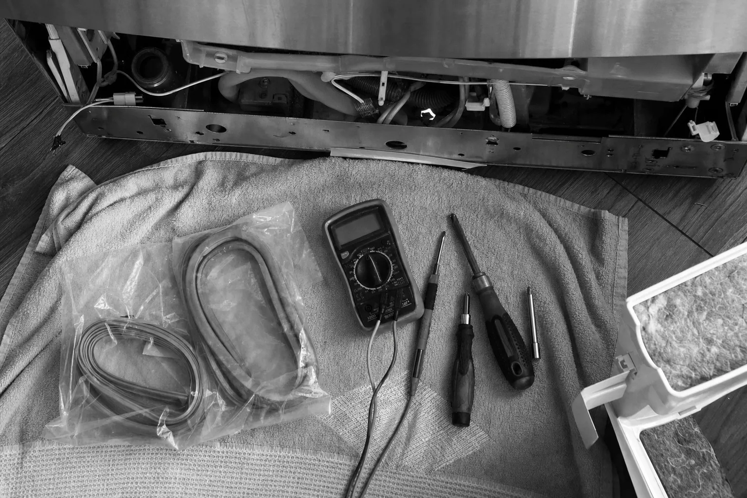 Black-and-white flat-lay of bagged replacement gaskets, a multimeter and screwdrivers on a towel before an open base